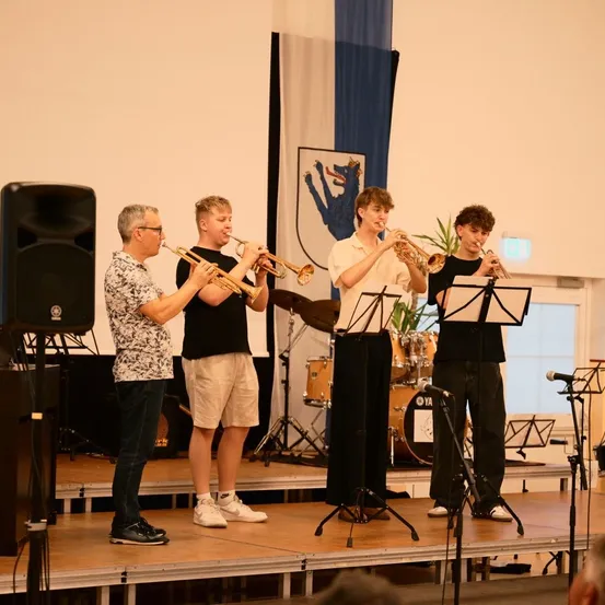 Vier Musiker stehen auf der Bühne und spielen Trompeten. Ein Mann steht links und drei junge Männer rechts. Hinter ihnen befinden sich Notenständer, ein Lautsprecher und ein Klavier.