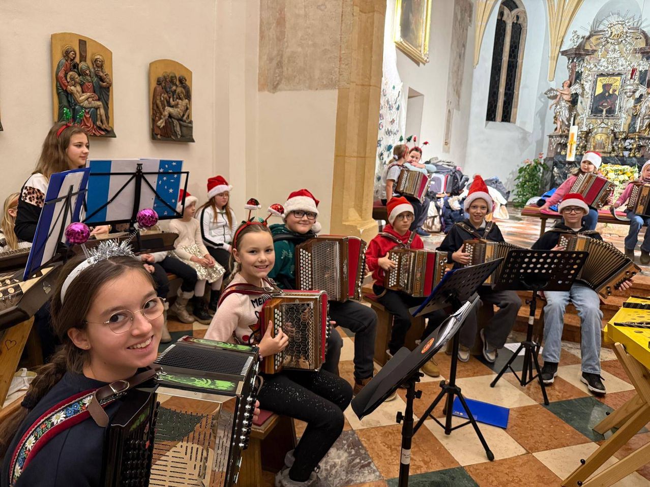 Eine Gruppe von Kindern spielt Akkordeons in einer Kirche, geschmückt mit Weihnachtsmützen und festlichen Dekorationen. Sie sitzen auf Bänken, während einige stehen, umgeben von Notenblättern. Die Kirche ist mit religiösen Gemälden an der Wand verziert.