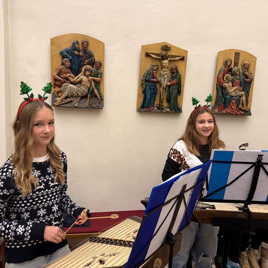 Zwei Mädchen mit Weihnachtsbändchen spielen ein Saiteninstrument und lesen Noten vor drei religiösen Gemälden.