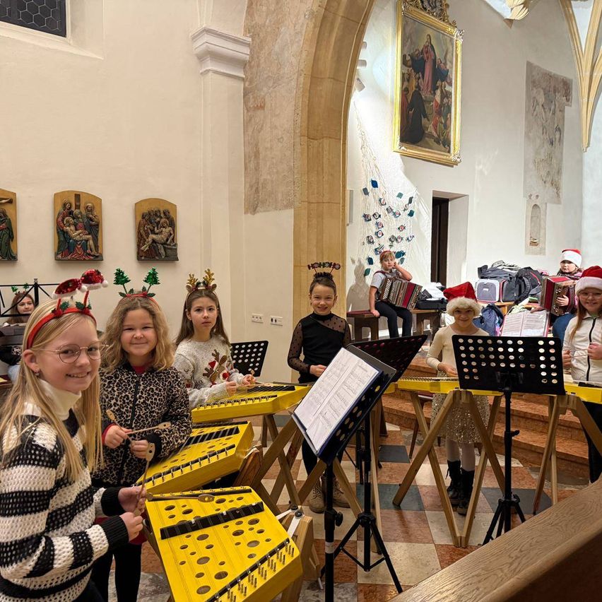 Eine Gruppe von Kindern mit Weihnachtskopfbändern und Brillen spielt ein Xylophon in einer Kirche. Einige lesen Musik von Notenständern.