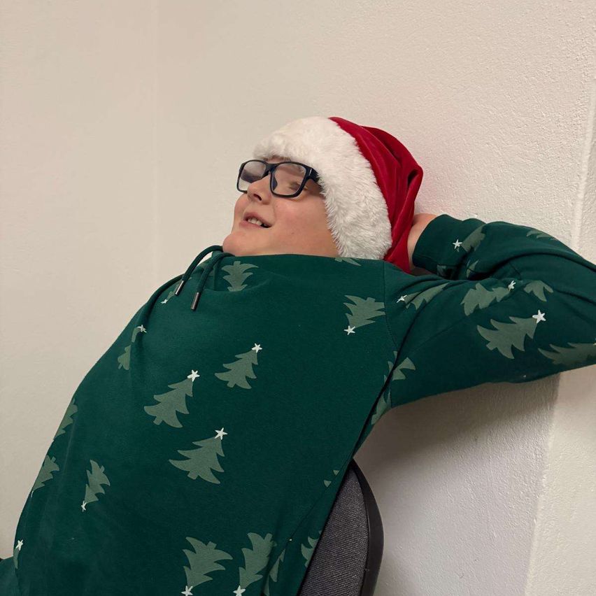 Eine Person mit einem Weihnachtsmannhut und Brille lehnt sich gegen eine weiße Wand und trägt ein grünes Sweatshirt mit Weihnachtsbaummuster.