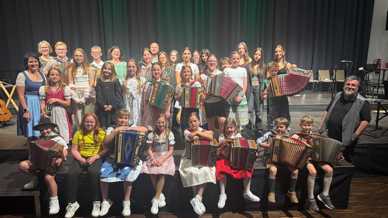 Group of children and adults holding accordions, smiling, standing on stage, green curtain in background, one child with drumstick.