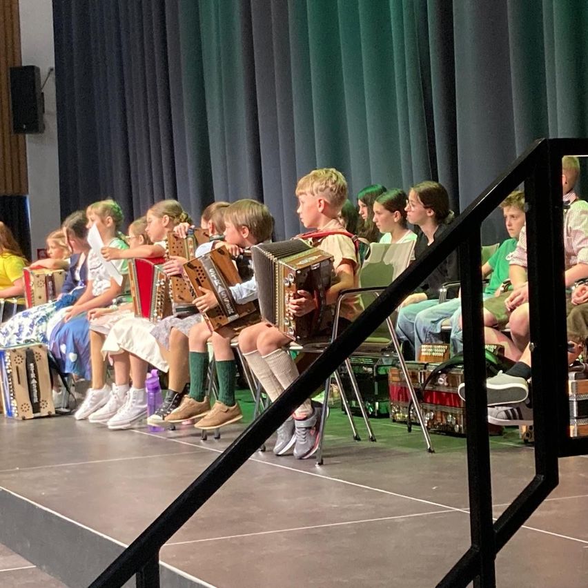 Several children play accordions on stage. They are seated and appear to be performing. Behind them are green curtains.