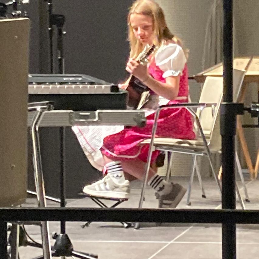 A girl sits on a stool with a guitar, wearing a pink dress, looking down, and playing a piano. She has sneakers on and the room is dimly lit.