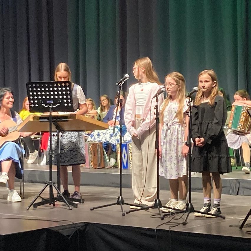 On a stage, a group of young girls perform a musical act. One holds a guitar, others play accordion. Audience members sit in the background.