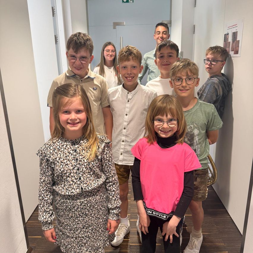 A group of young children in glasses, smiling for a photo. Some wear casual clothes, while others have more formal attire. They are standing in a corridor with a wooden floor and white walls.