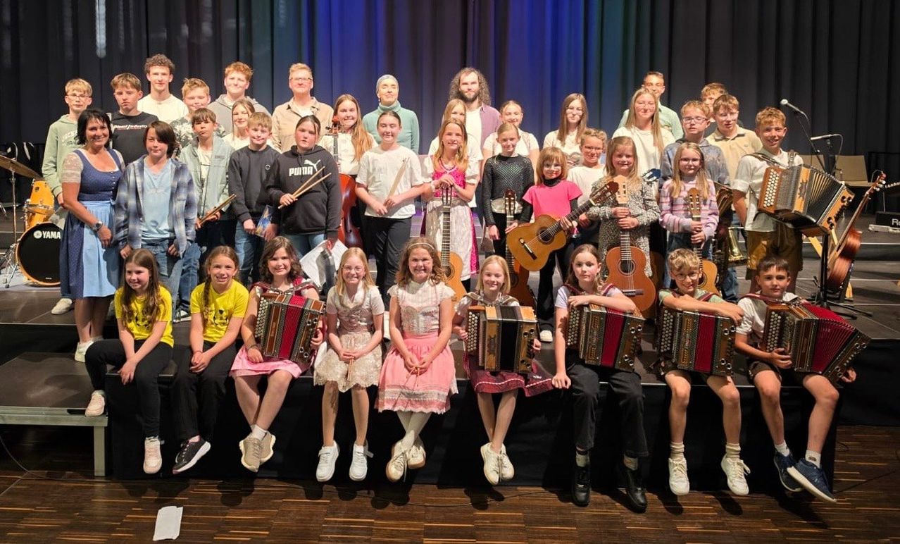 A group of children and young adults are posing for a photo on a stage with musical instruments. Some of them are holding guitars, while others have accordions. They are smiling and seem to be happy.