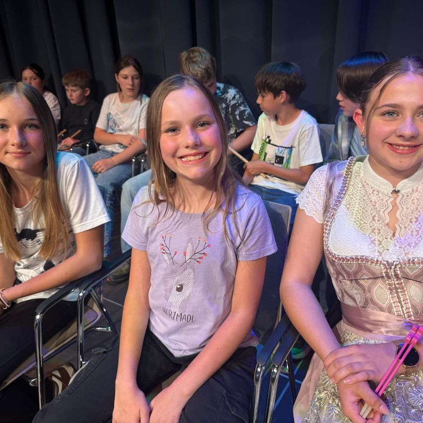 A group of children sitting in chairs with some holding drumsticks. A girl in a purple shirt with a deer design and a girl in a white dress with lace are smiling.