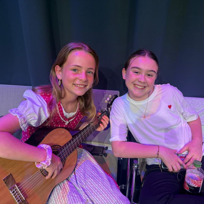 Two young girls smiling and posing for a photo. One holds a guitar, the other has a drink bottle. They sit on chairs with a purple backdrop.