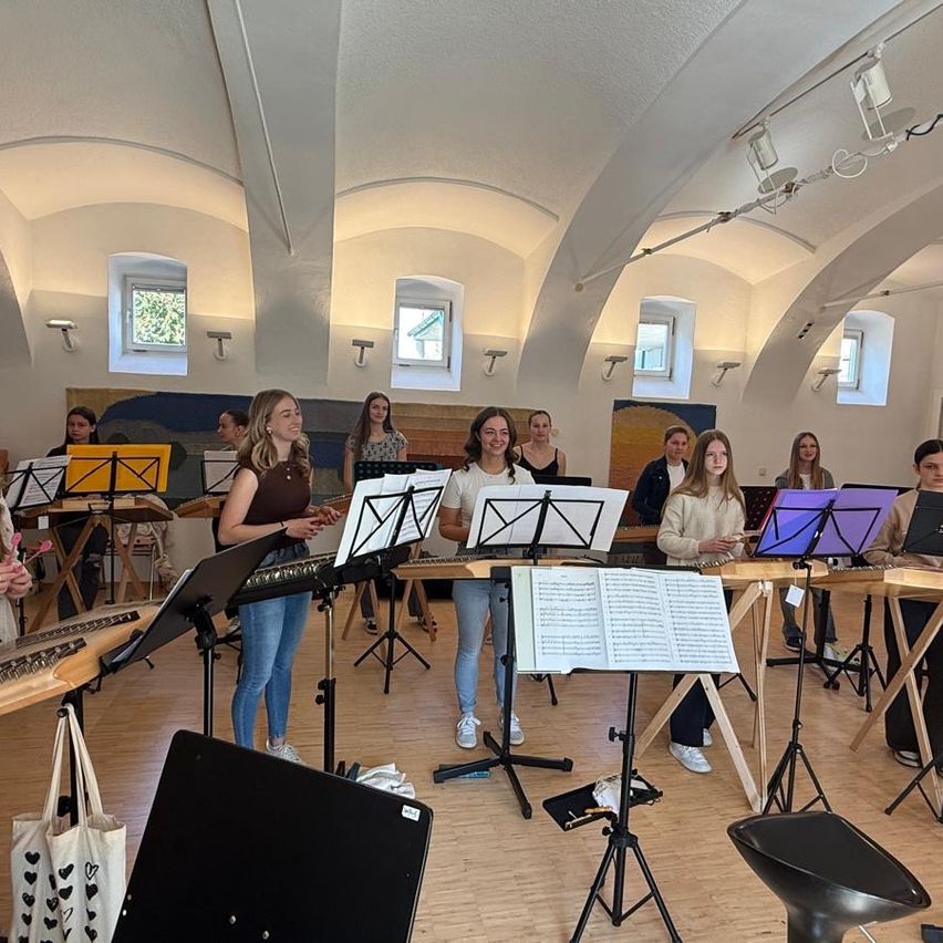 Eine Gruppe junger Frauen übt in einem Musikraum und spielt verschiedene Instrumente wie Xylophon und Harfe. Sie stehen vor Notenständern mit Musikblättern.