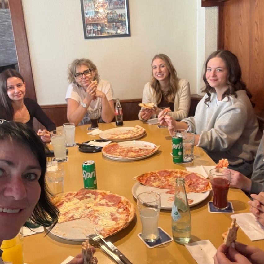 Eine Gruppe von Menschen sitzt an einem Tisch und isst Pizza und trinkt verschiedene Getränke. Eine Frau mit Brille hält ein Pizzastück. Der Tisch ist voll mit Pizzaplatten, Gläsern und Dosen.