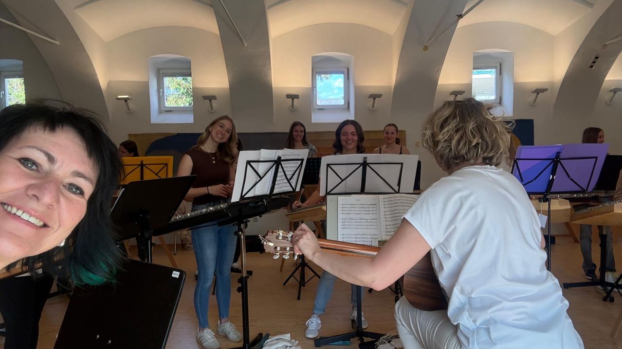 Eine Gruppe von Frauen übt in einem Musikraum. Sie schauen Notenständer an und eine spielt Gitarre.