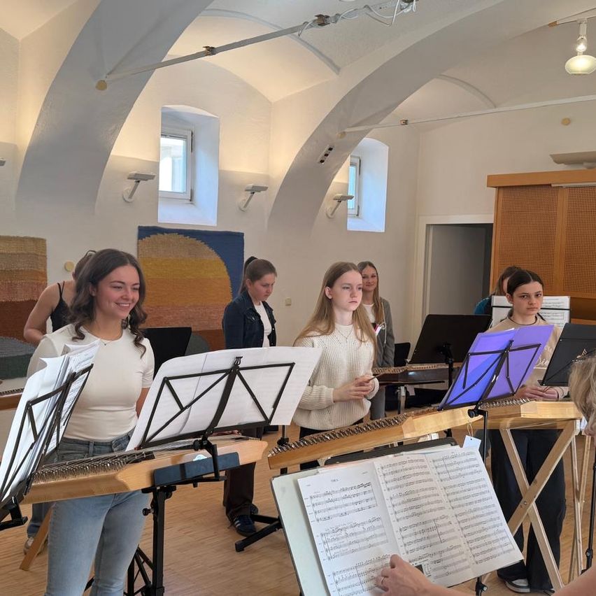 Eine Gruppe von Mädchen übt mit Saiteninstrumenten, darunter Harfen, in einem Raum mit Holzböden und gewölbten Decken. Sie sehen Noten auf einem Notenständer.