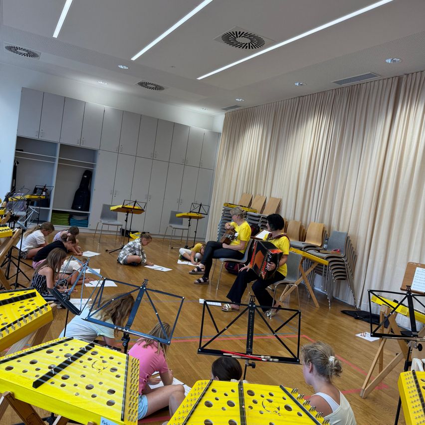 A group of children and adults are in a music room, practicing with various instruments. Some are seated, others are on the floor, and one person is playing an accordion.