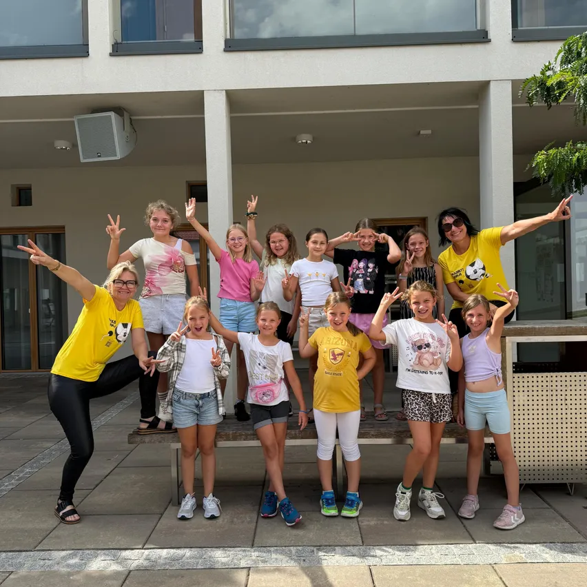 Eine Gruppe junger Mädchen und zwei erwachsene Frauen posieren für ein Foto vor einem Gebäude. Sie tragen alle Freizeitkleidung und lächeln.