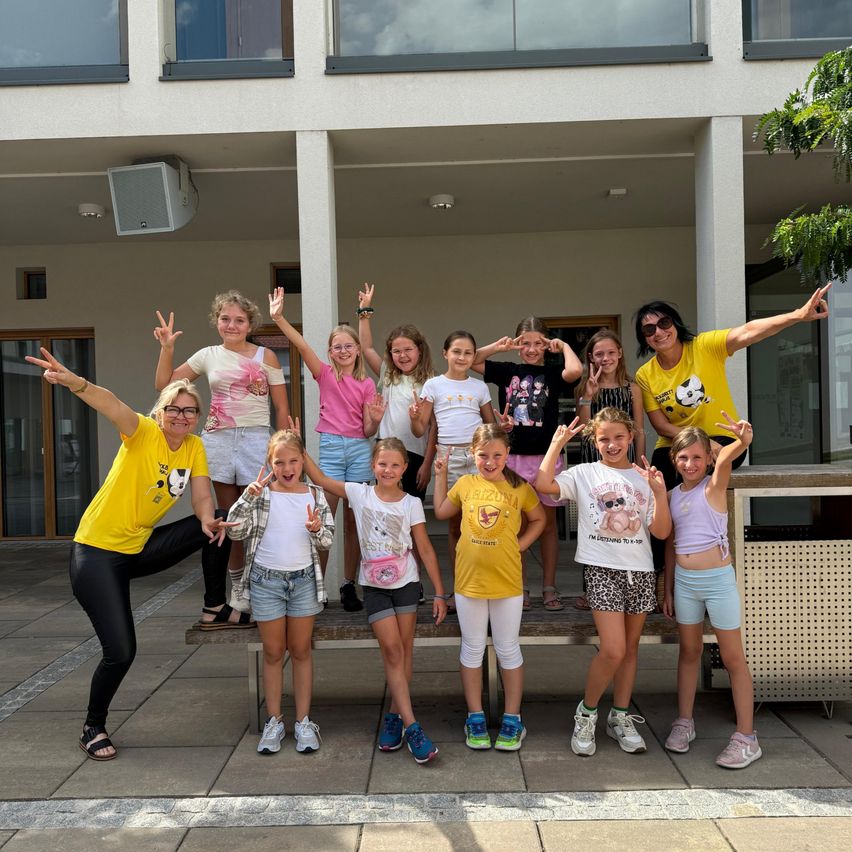A group of young girls and two adult women are posing for a photo in front of a building. They are all wearing casual clothes and smiling.