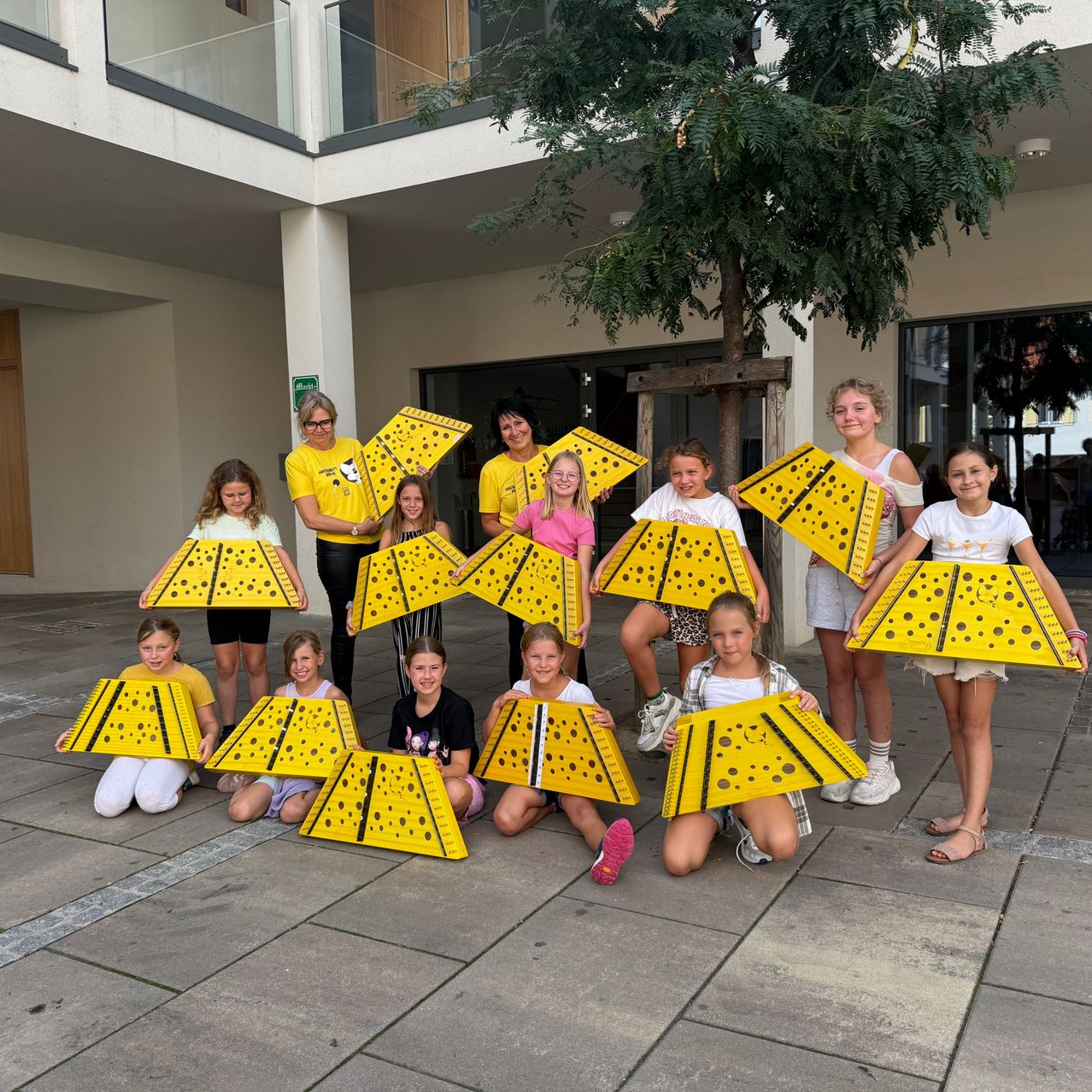 A group of children and adults pose for a photo, holding up yellow triangular boards with black dots. They are standing in a row in front of a building.