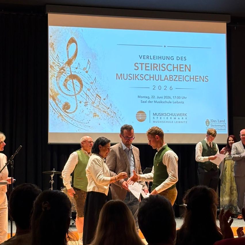 Eine Zeremonie an der Musikschule Leibnitz. Eine Frau überreicht einem Studenten ein Zertifikat vor einem Publikum. Ein Bildschirm hinter ihnen zeigt Datum und Veranstaltungsname.