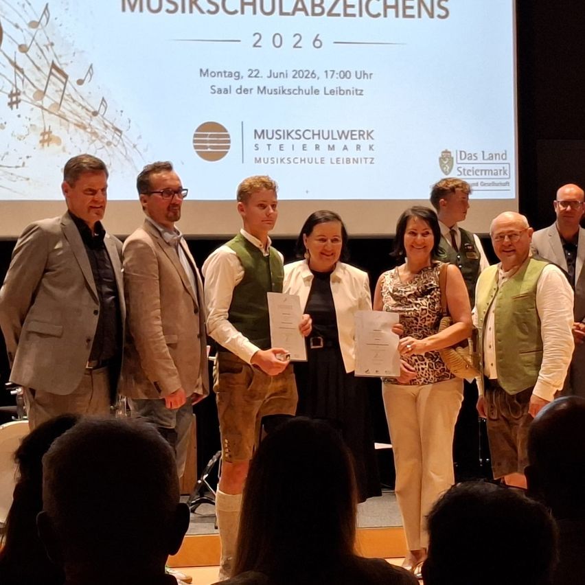 Mehrere Personen stehen auf der Bühne und halten Urkunden in den Händen. Ein Bildschirm hinter ihnen zeigt Datum und Namen einer Veranstaltung. Zuschauer sind im Vordergrund zu sehen.