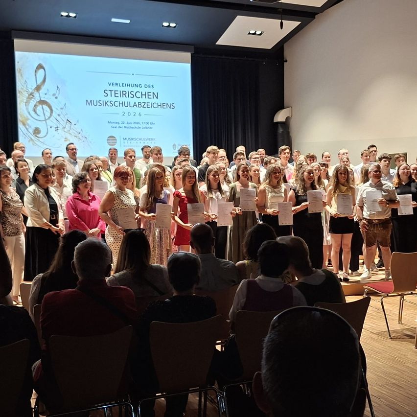 Eine Gruppe von Menschen steht mit Urkunden auf der Bühne, vor ihnen sitzen Zuschauer. Eine Leinwand hinter ihnen zeigt eine Musikpartitur.