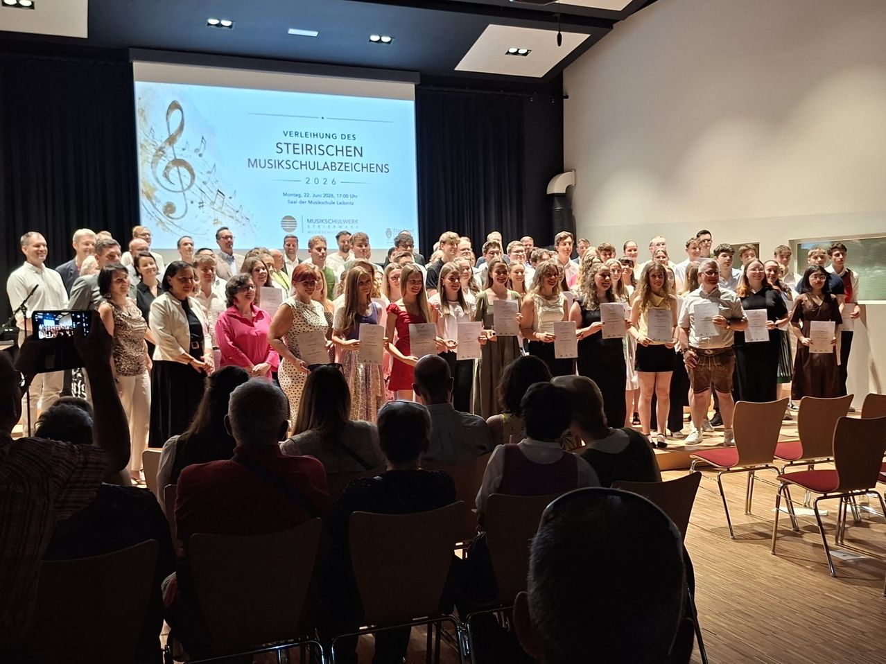 Eine Gruppe von Menschen steht mit Urkunden auf der Bühne, vor ihnen sitzen Zuschauer. Eine Leinwand hinter ihnen zeigt eine Musikpartitur.