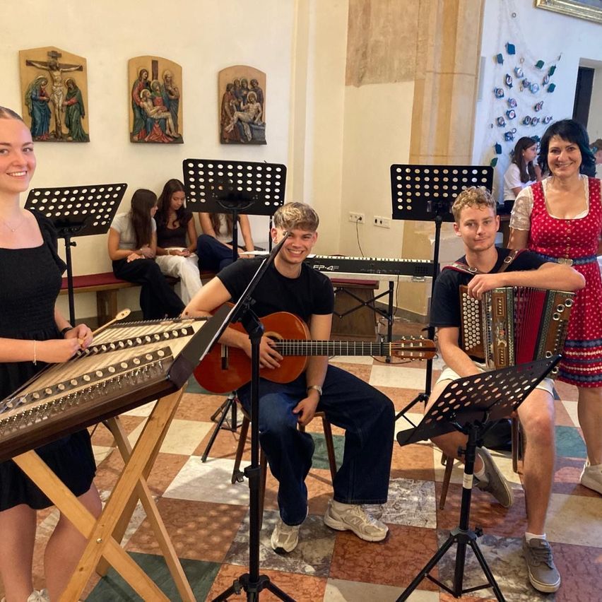 Eine Gruppe junger Musiker spielt in einer Kirche mit religiösen Gemälden an der Wand. Sie sitzen auf Stühlen und spielen verschiedene Instrumente.