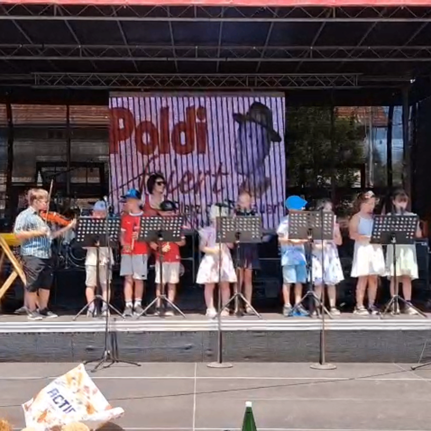 Eine Gruppe von Kindern auf der Bühne spielt Instrumente. Dahinter befindet sich eine große Leinwand mit dem Wort 'Paldi'.