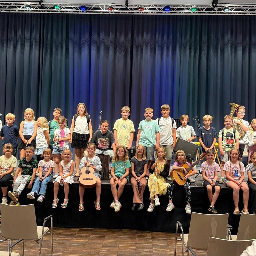 Eine Gruppe von Kindern, einige stehend, einige sitzend, sind mit Musikinstrumenten auf der Bühne. Sie tragen Freizeitkleidung und scheinen sich in einer Klassenzimmerumgebung zu befinden.