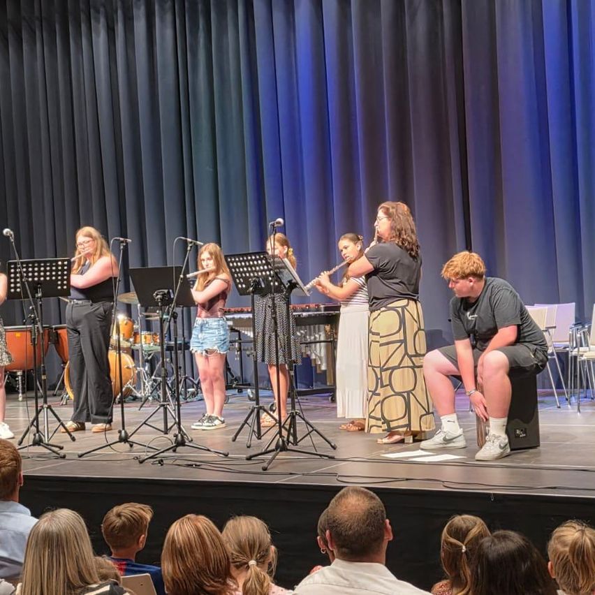 Eine Gruppe von Musikern tritt auf einer Bühne mit einem blauen Vorhang auf. Sie spielen Flöten, ein Schlagzeug und andere Instrumente. Ein Publikum beobachtet sie.