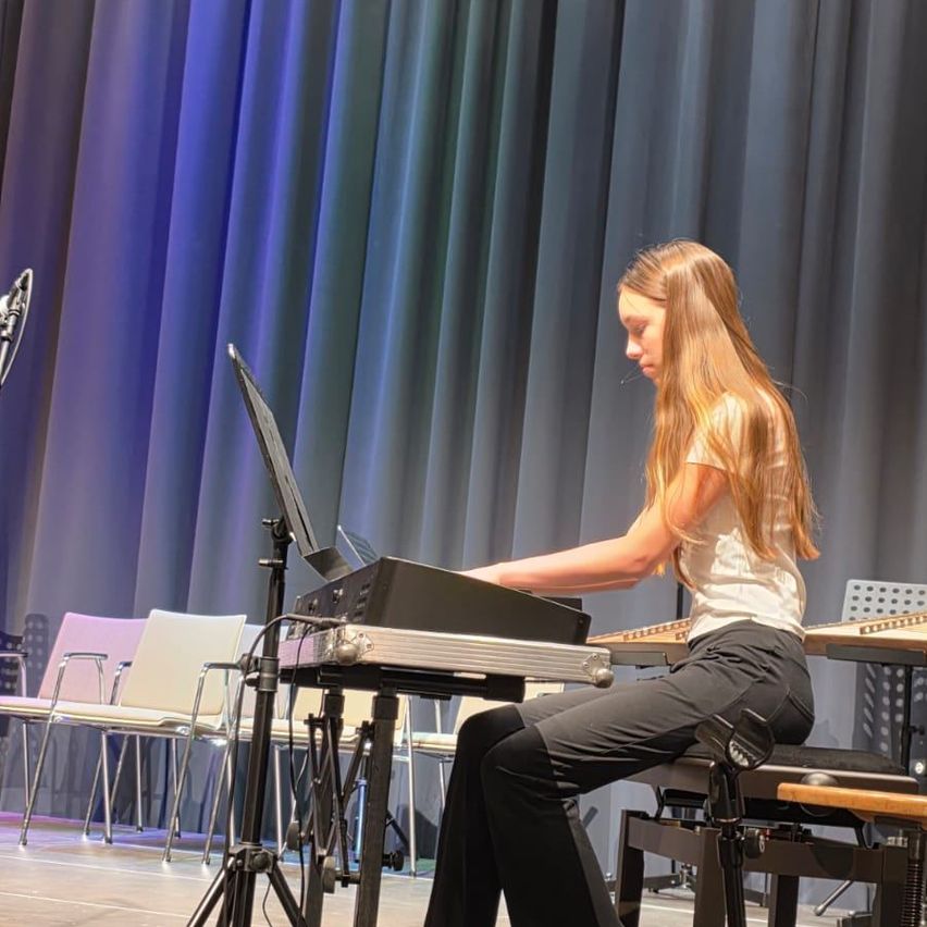 Ein Mädchen mit langen Haaren spielt eine Tastatur auf der Bühne, mit einem Mikrofon davor und Stühlen hinter einem Vorhang.