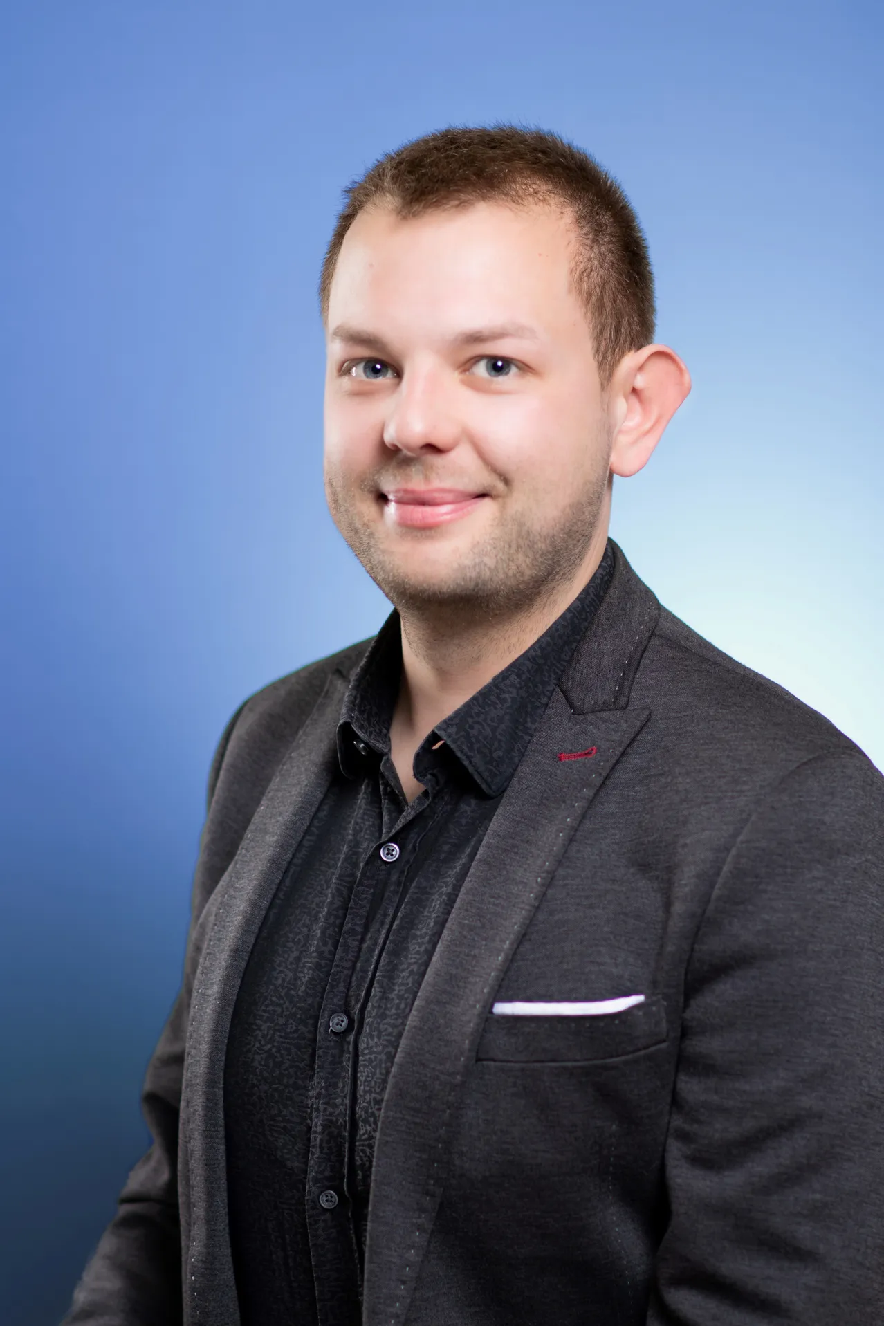 Ein Mann mit kurzen braunen Haaren lächelt und schaut in die Kamera. Er trägt ein schwarzes Hemd und einen grauen Blazer.