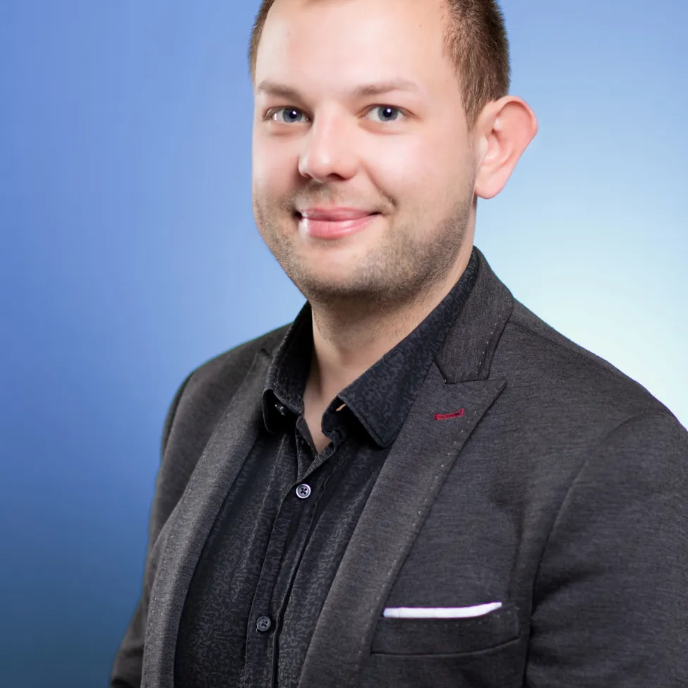 Ein Mann mit kurzen braunen Haaren lächelt und schaut in die Kamera. Er trägt ein schwarzes Hemd und einen grauen Blazer.