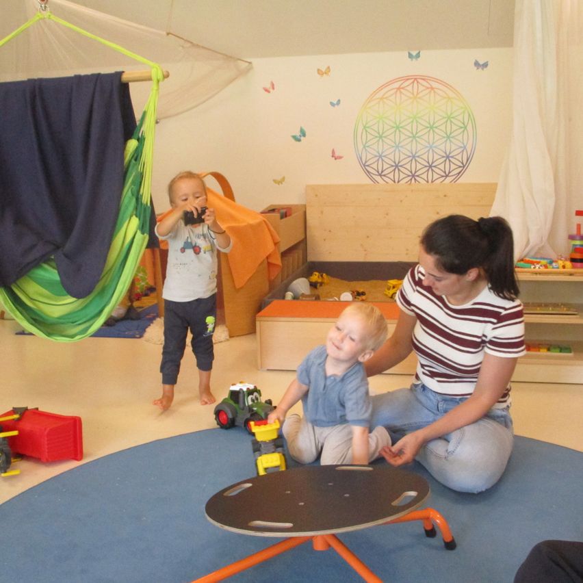 Zwei Kinder und eine Frau sind in einem Raum mit Spielzeug, einer Hängematte und einem Blumenwandbild. Die Frau sitzt mit einem Kind auf einem kleinen Tisch.