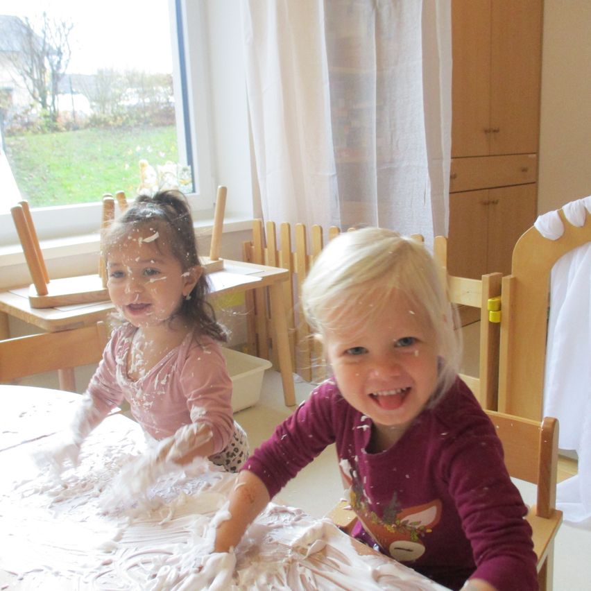 Zwei kleine Mädchen sitzen an einem Tisch, der mit Schaum bedeckt ist. Ein Mädchen spielt mit dem Schaum, während das andere lächelt. Hinter ihnen stehen hölzerne Stühle, ein Fenster mit Vorhängen und ein Rasen.