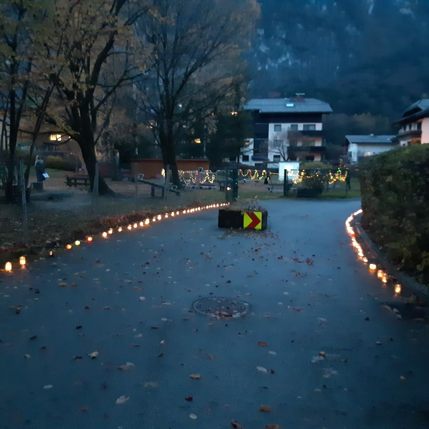 A pathway illuminated by candles and a road sign, surrounded by trees and buildings, in a mountainous area.