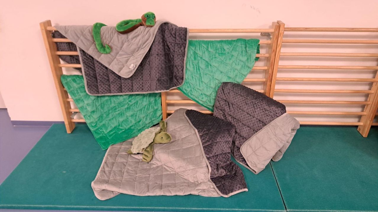 Verschiedene Gewichtsdecken, einschließlich Grün, Grau und Teal, sind auf einem Holzrahmen ausgestellt. Ein grünes Schildkröten-Plüschtier ruht auf einer der Decken.