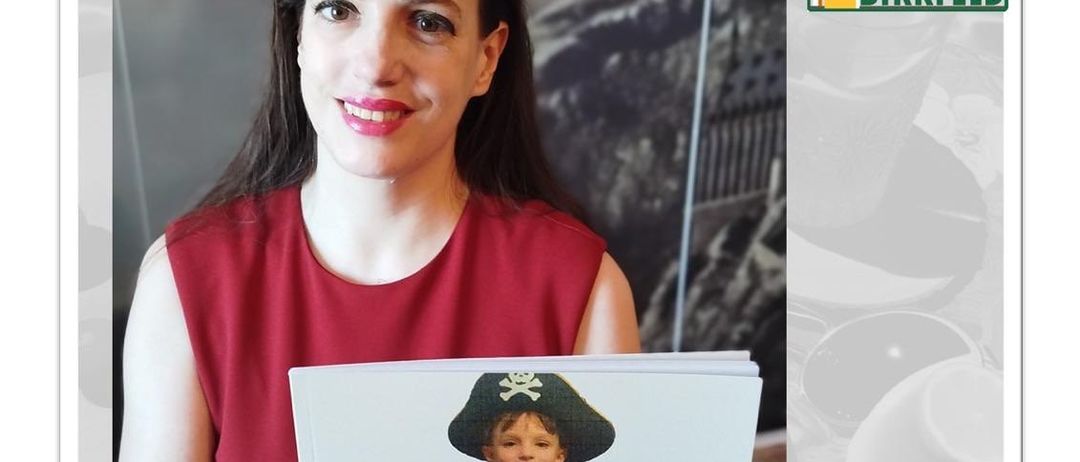 Eine Frau hält ein Buch mit dem Titel 'Markus, Marcus und Mark der weisse Pirat' mit einem Kinderbild auf dem Cover und lächelt für ein Foto. Der Hintergrund enthält ein Bibliothekslogo und ein verschwommenes Bild eines Berges.