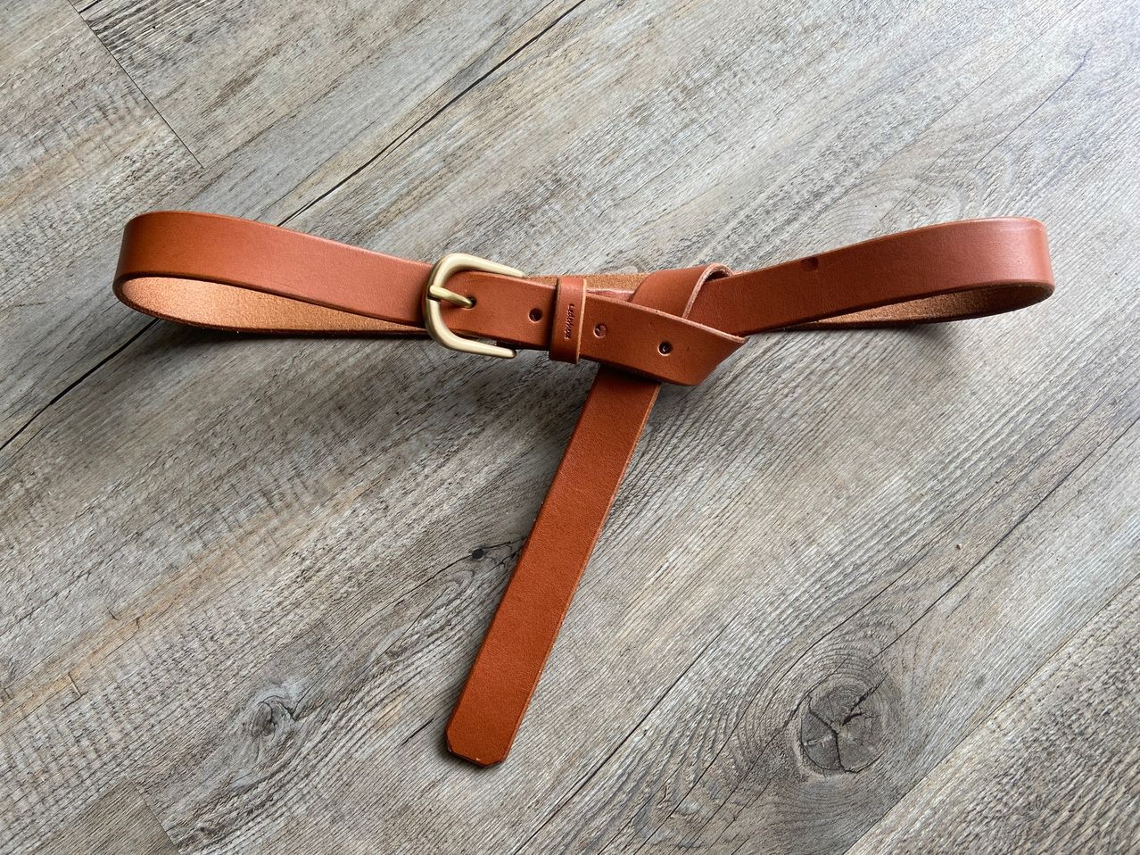 Ein brauner Ledergürtel mit einer goldenen Schnalle liegt auf einer hölzernen Oberfläche. Der Gürtel hat ein schlichtes Design mit einer glatten Textur.