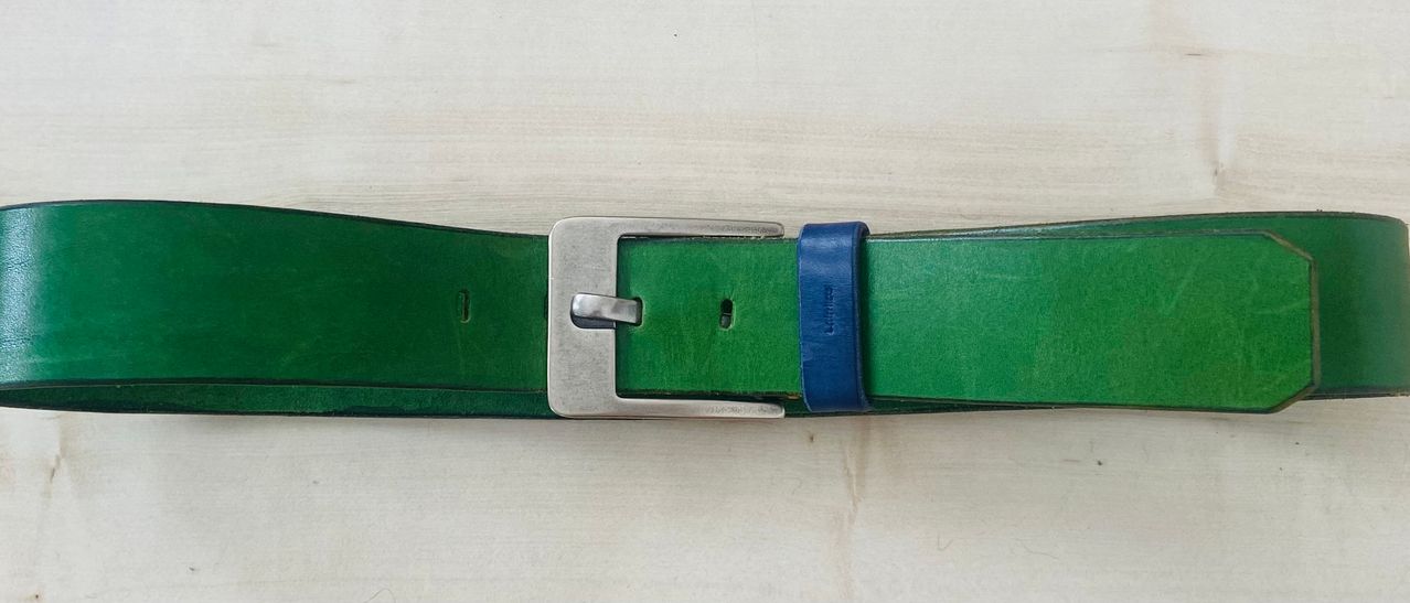 Ein grüner Ledergürtel mit einer silbernen Schnalle und einem blauen Lederstück an der Seite. Er hat ein klares Aussehen und ein einfaches Design.