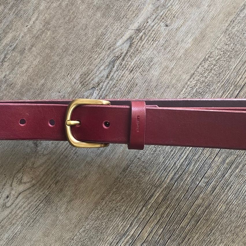 Ein roter Ledergürtel mit einer goldenen Schnalle liegt flach auf einer Holzoberfläche. Der Gürtel hat ein einfaches Design mit kleinen Löchern für Gürtelschlaufen.