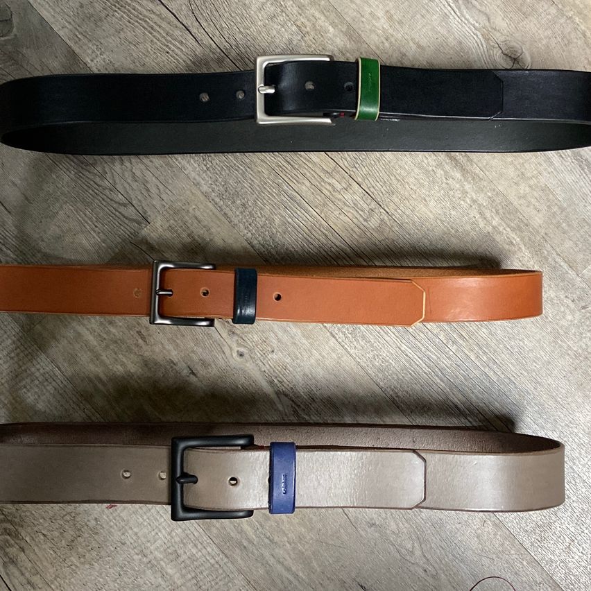 Drei Ledergürtel in verschiedenen Farben und Stilen liegen auf einer Holzoberfläche. Der obere Gürtel ist schwarz mit einer silbernen Schnalle und einem grünen Streifen. Der mittlere Gürtel ist braun mit einer silbernen Schnalle und einem schwarzen Streifen. Der untere Gürtel ist braun mit einer schwarzen Schnalle und einem blauen Streifen.