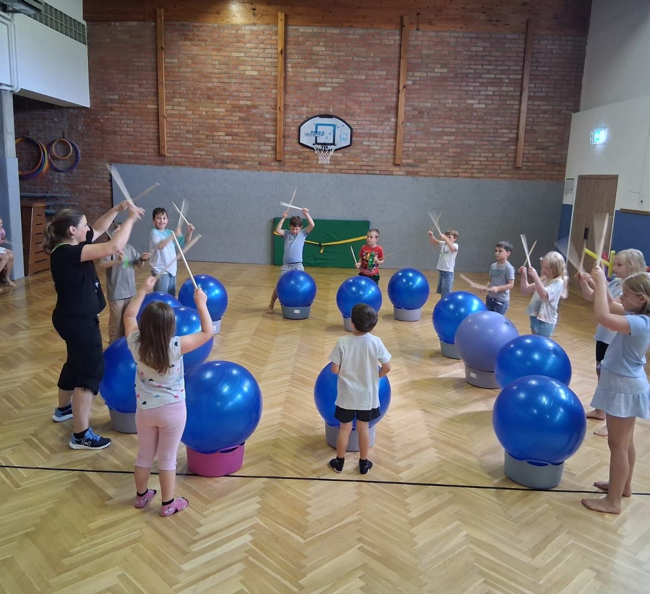 Mehrere Kinder und Erwachsene sind in einer Turnhalle und spielen mit einer Trommel und Stöcken. Sie stehen um blaue Übungsbälle herum. Die Turnhalle hat Ziegelwände und einen Basketballkorb.
