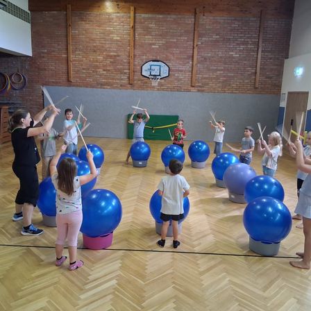 Mehrere Kinder und Erwachsene sind in einer Turnhalle und spielen mit einer Trommel und Stöcken. Sie stehen um blaue Übungsbälle herum. Die Turnhalle hat Ziegelwände und einen Basketballkorb.