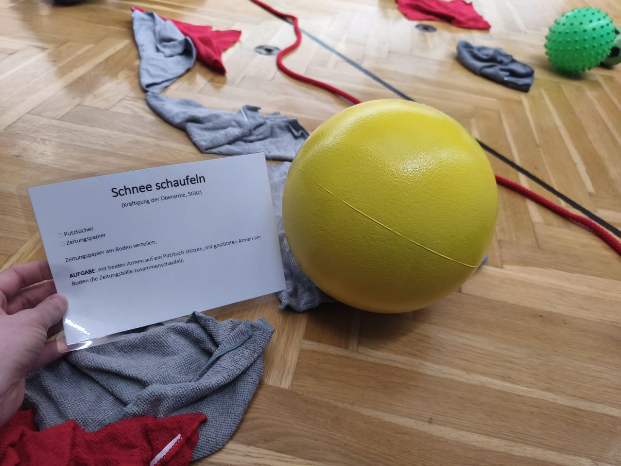 Ein gelber Übungsball mit Anweisungen auf einer Karte für eine Schulterübung namens 'Schnee schaufeln' liegt auf einem Holzboden. Verschiedene Kleidungsstücke sind herum verstreut.