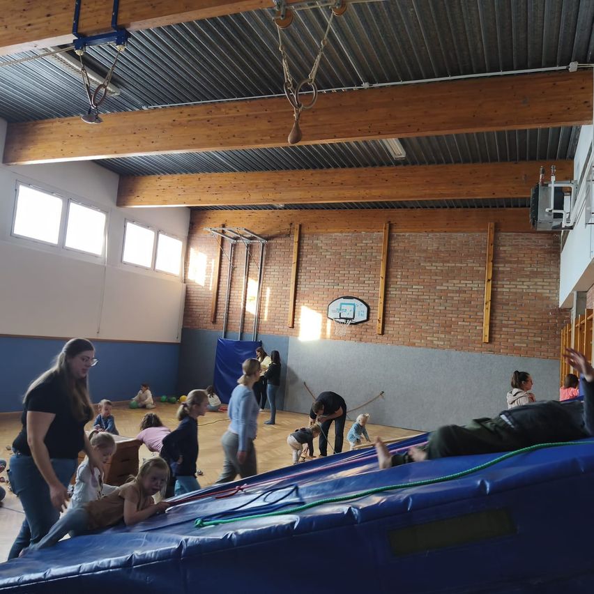 Innengymnasium mit Kindern, die auf einem blauen aufblasbaren Schlitten spielen, Erwachsene, die beaufsichtigen und herumstehen, Ziegelwände, ein Basketballkorb und Holzbalken an der Decke.