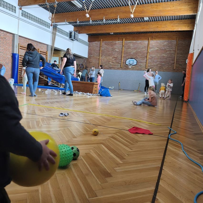 Sporthalle mit Holzboden, Menschen stehen, sitzen und spielen mit Bällen, einer Rutsche und einem Basketballkorb.
