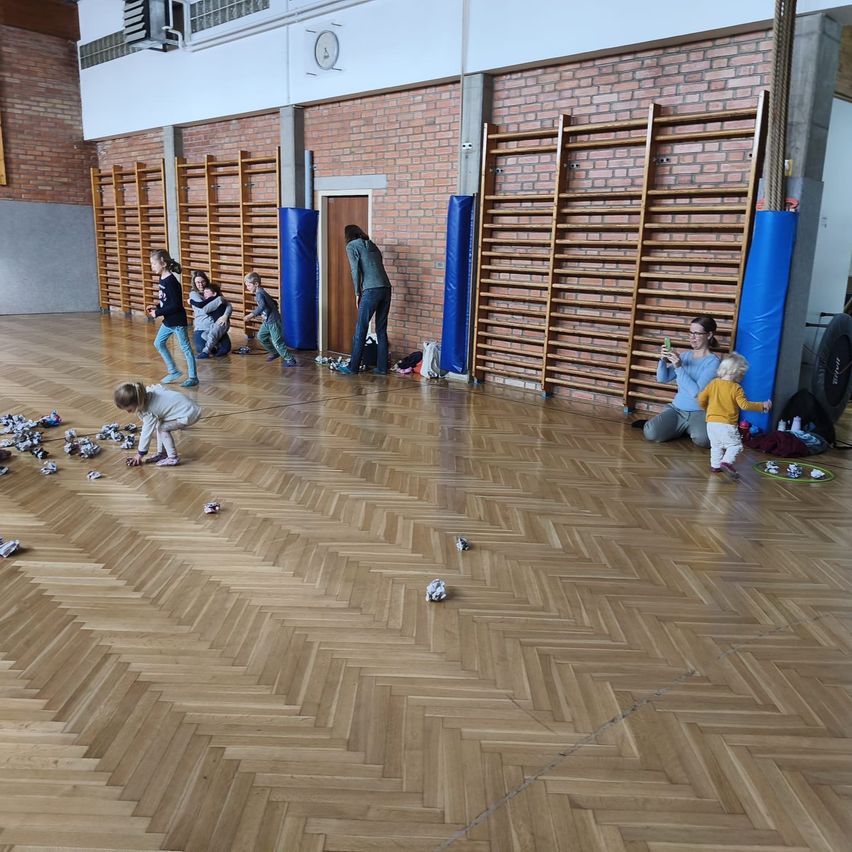 In einer Turnhalle spielen Kinder und Erwachsene mit kleinen Objekten auf einem Holzboden. Ziegelwände und Holzbalken sind im Hintergrund sichtbar.