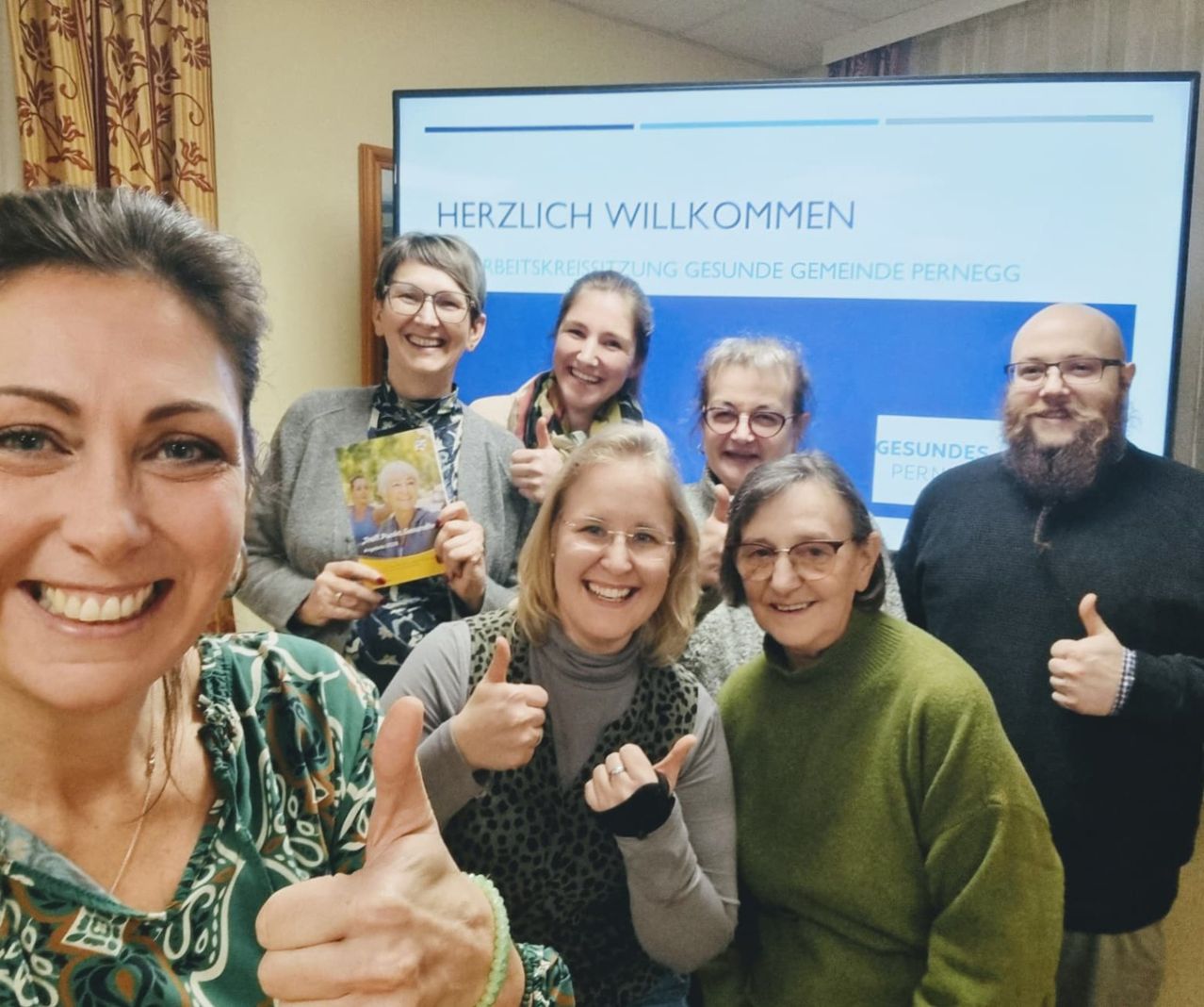 Eine Gruppe lächelnder Erwachsener steht vor einem Schild mit Herzlich Willkommen. Sie halten ein Buch und zeigen Daumen hoch.