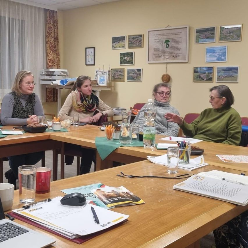 Eine Gruppe von Frauen sitzt um einen Tisch in einem Raum. Sie haben Snacks und Getränke vor sich. Der Tisch hat Papiere, Stifte und einen Laptop. Der Raum hat eine gelbe Wand mit mehreren Bilderrahmen.
