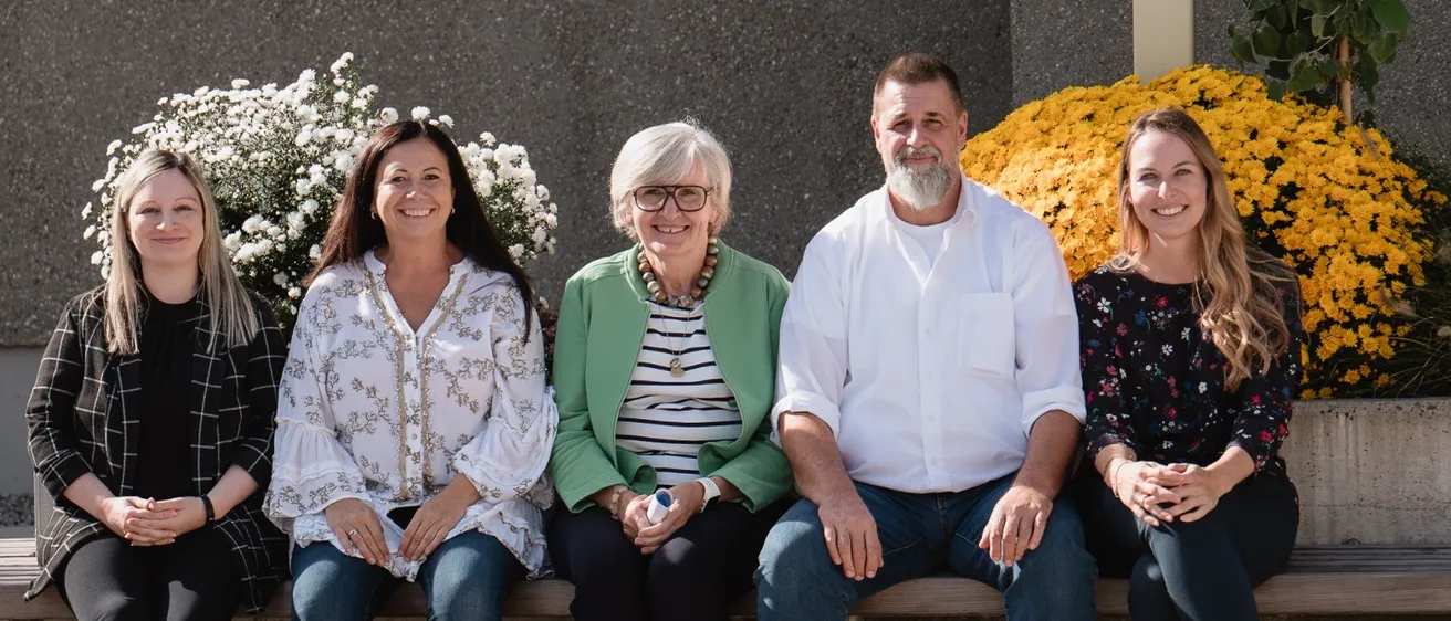 Fünf Personen sitzen auf einer Bank vor gelben und weißen Blumen. Sie lächeln und scheinen für ein Foto zu posieren.